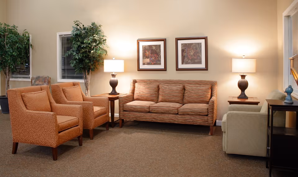 A cozy living room area with a striped sofa, two patterned armchairs, and a green armchair. There are two table lamps on wooden side tables, two framed floral artworks on the wall, and two large potted plants near the windows.