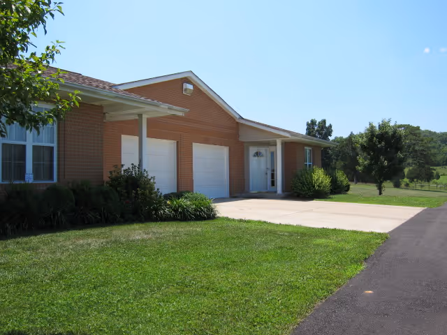 Exterior view of a single-story brick building with two white garage doors and a white front door. The building is surrounded by green grass, bushes, and trees under a clear blue sky.