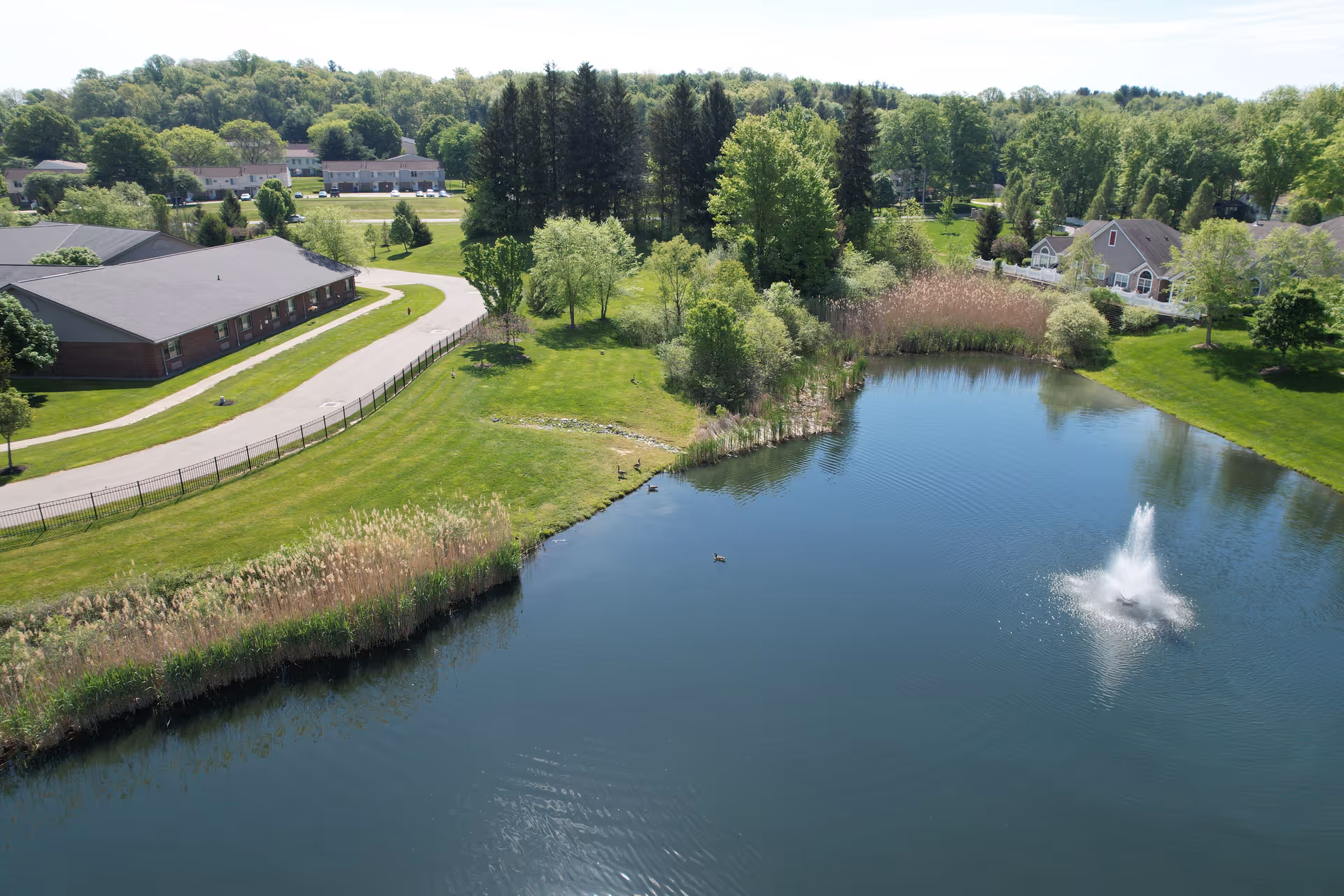 Aerial view of a senior living campus with a pond and fountain, surrounding lawns, trees, and adjacent buildings.