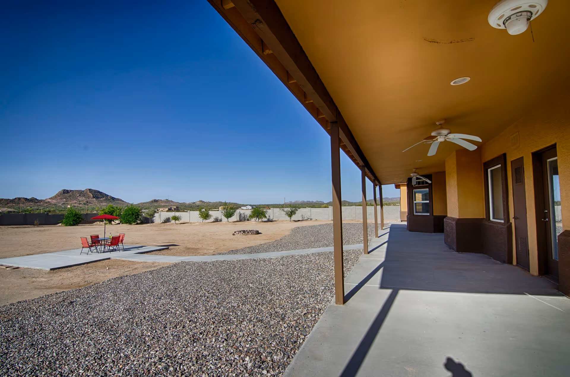 A covered outdoor patio area with ceiling fans and multiple doors leading inside. The patio overlooks a spacious yard with gravel and dirt ground, a concrete pad with a red umbrella and chairs, some small trees, and mountains in the distance under a clear blue sky.