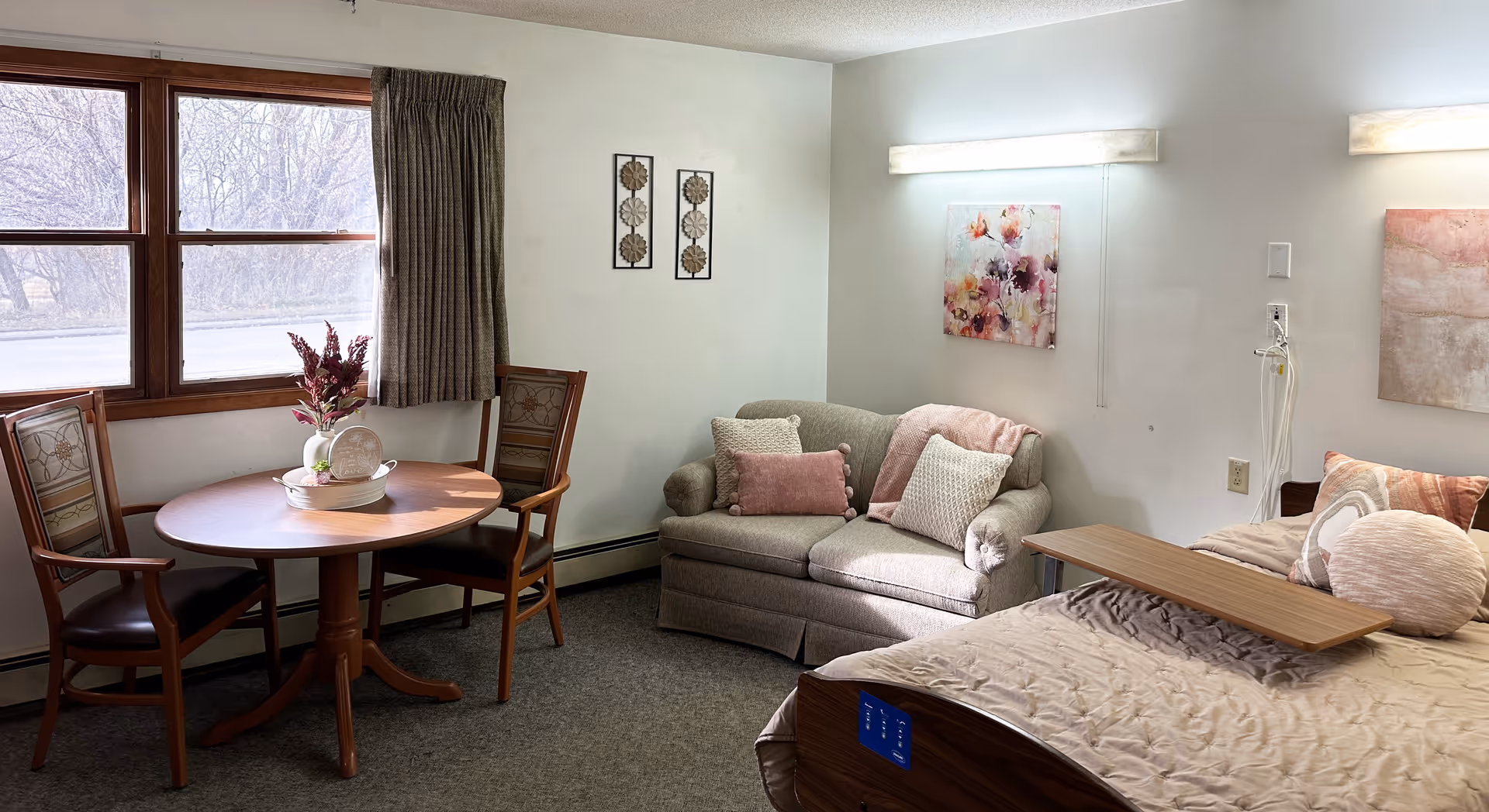 A cozy senior living room featuring a round wooden table with two chairs near a window with curtains. There is a small gray loveseat with decorative pillows and a pink throw blanket. Next to the loveseat is a bed with a beige quilt, multiple pillows, and a wooden overbed table. The walls are decorated with floral artwork and two wall-mounted lights.
