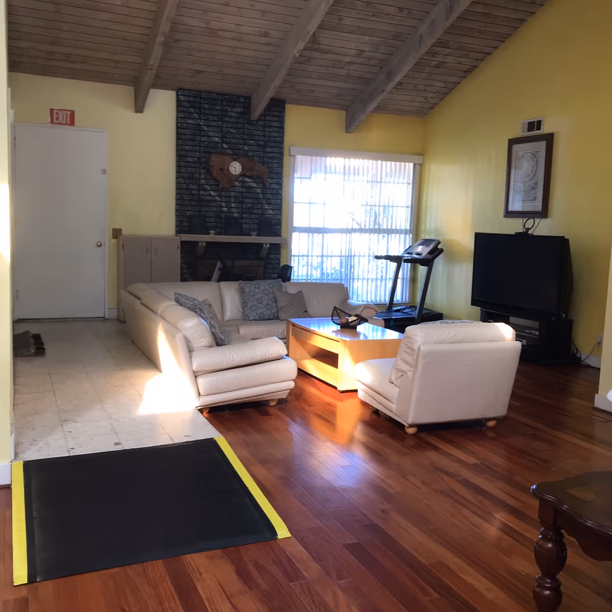 A cozy living room with yellow walls and a wooden vaulted ceiling. The room features a beige leather sectional sofa and a matching armchair arranged around a wooden coffee table. There is a treadmill near a large window with vertical blinds, a TV on a stand against the wall, and a fireplace with a decorative clock above it. The floor is a combination of wood and tile with a black ramp connecting the two areas.