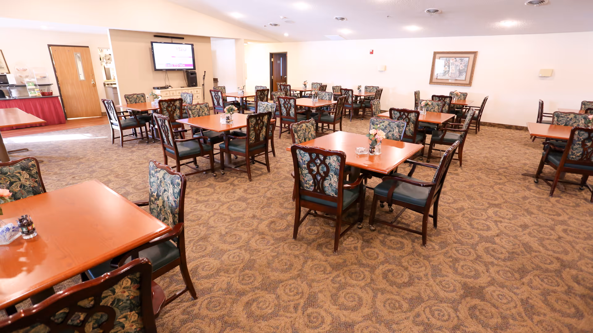 A dining room with multiple wooden tables and chairs with floral upholstery. Each table has a small flower arrangement and condiments. There is a television mounted on the wall and a door leading to another room. The carpet has a patterned design and the walls are light-colored with a framed picture hanging on one side.