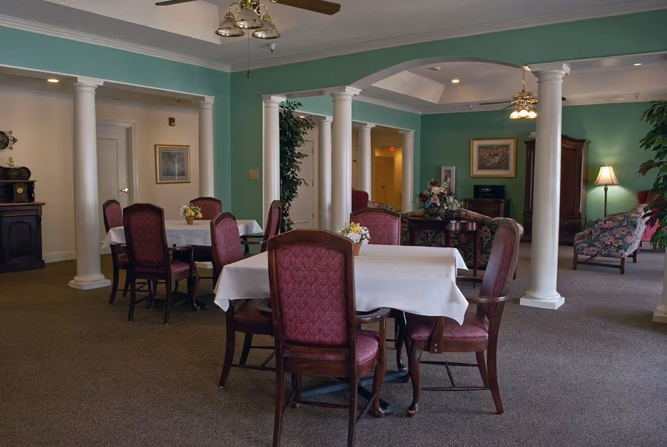 Interior view of a senior living facility dining and lounge area with tables covered in white tablecloths, surrounded by wooden chairs with red upholstery. The room features green walls, white columns, carpeted floor, ceiling fans with lights, and floral arrangements on the tables. In the background, there is a seating area with floral-patterned armchairs, a floor lamp, and framed artwork on the walls.
