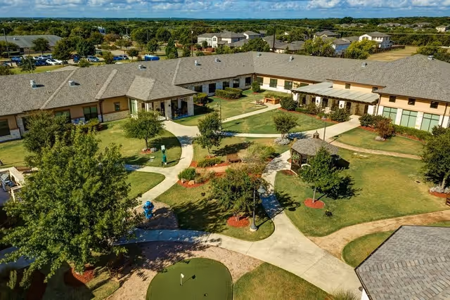 Aerial view of a senior living facility courtyard with well-maintained green lawns, walking paths, trees, benches, a small gazebo, and a putting green. The surrounding single-story buildings have beige walls and gray shingled roofs under a partly cloudy sky.