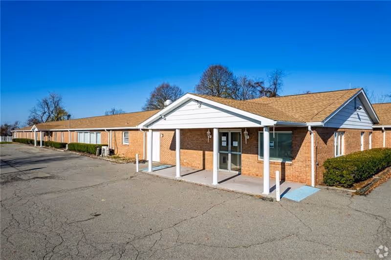 Single-story brick senior living facility with a covered main entrance and a large paved parking area under a clear blue sky.