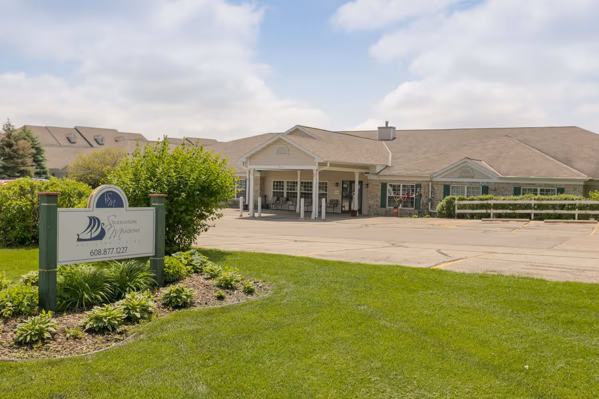 Front exterior of Stoughton Meadows Assisted Living building with a lawn, entrance canopy, parking area, and a sign in the foreground.