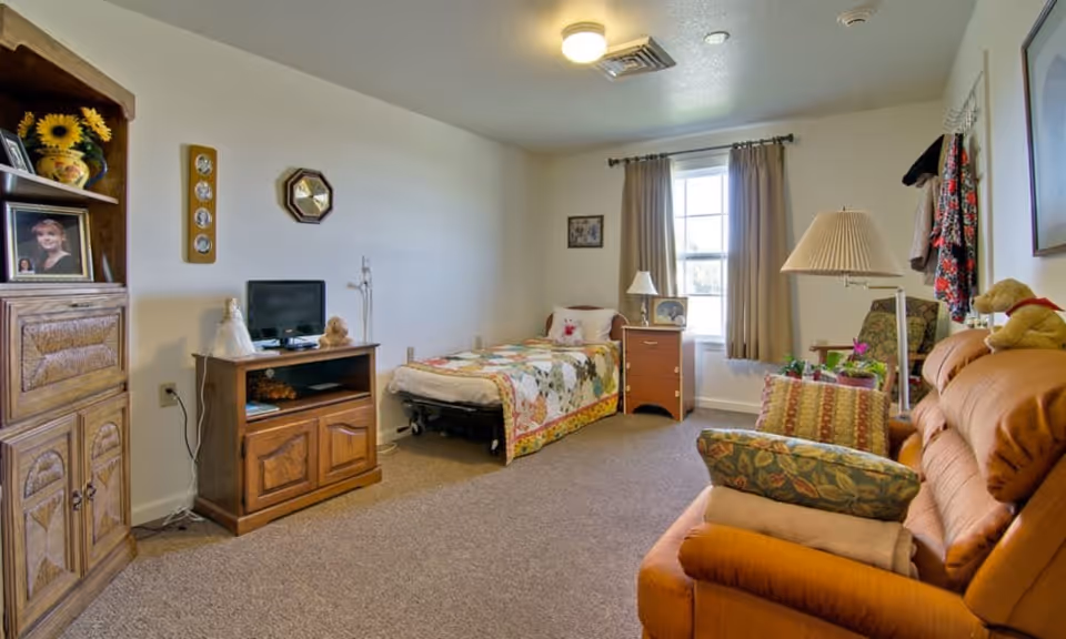 A cozy senior living bedroom featuring a single bed with a colorful quilt, a wooden nightstand with a lamp and framed photo, a wooden TV stand with a small television and decorative items, a wooden cabinet with framed photos and a vase with sunflowers, a comfortable orange sofa with patterned cushions, a floor lamp, and a window with beige curtains letting in natural light.