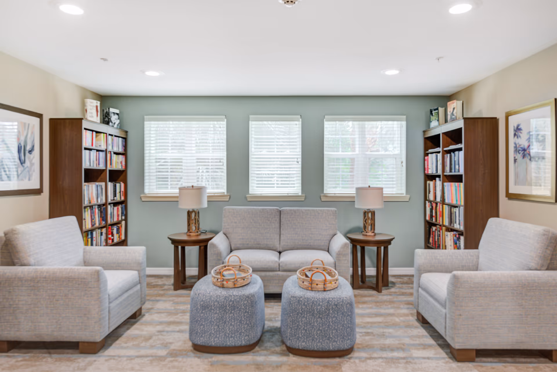 A cozy sitting area with two armchairs and a loveseat upholstered in light gray fabric. Two round wooden side tables with lamps are placed on either side of the loveseat. In front of the seating are two blue patterned ottomans with wicker baskets on top. Behind the seating area are three windows with white blinds, flanked by two tall wooden bookshelves filled with books. The walls are painted in a soft green and beige color, and framed artwork hangs on the side walls.