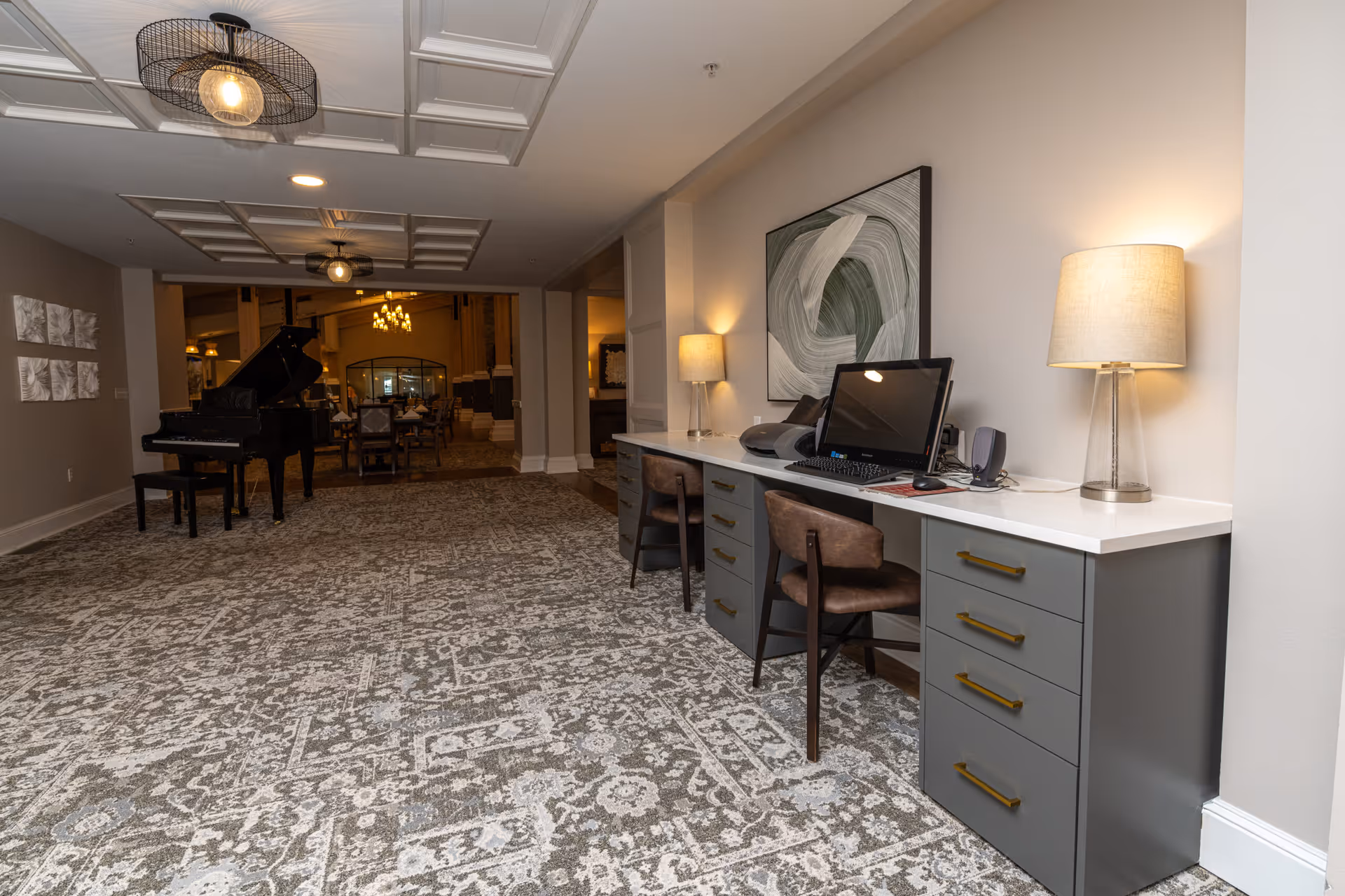 Interior view of a senior living facility hallway with a patterned carpet, a desk with two chairs, a computer, and two table lamps on the right side. In the background, there is a grand piano and a dining area with a chandelier.