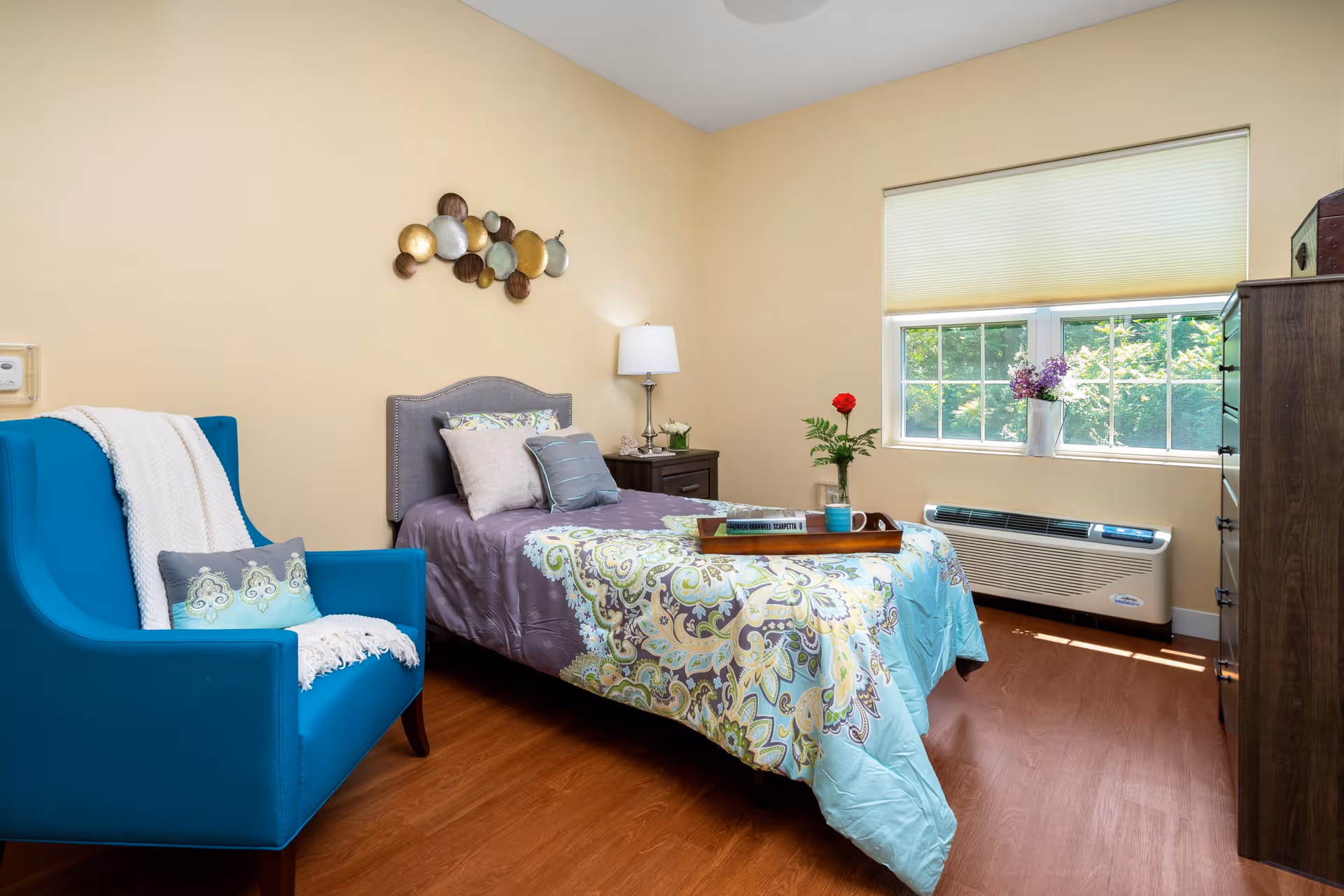 A cozy bedroom with a single bed covered in a patterned quilt and multiple pillows. Next to the bed is a dark wooden nightstand with a lamp and a small plant. A blue armchair with a white throw blanket and a decorative pillow is positioned to the left. A window with a beige blind lets in natural light, and a wooden dresser is on the right side of the room. The walls are painted light beige, and there is a decorative metal wall art above the bed.