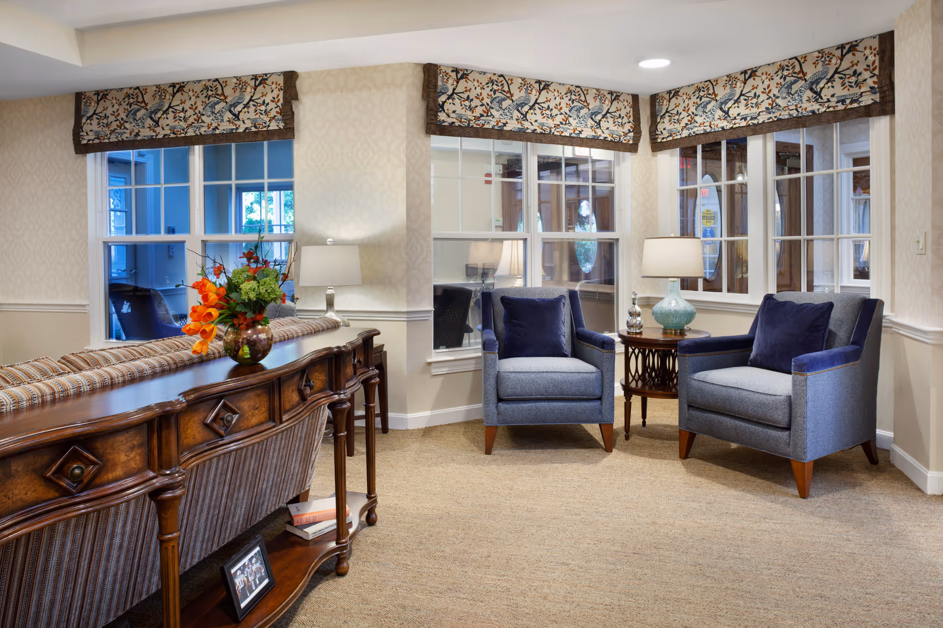 Comfortable sitting area with two blue armchairs, a side table with lamps, and a wooden console topped with a floral arrangement.