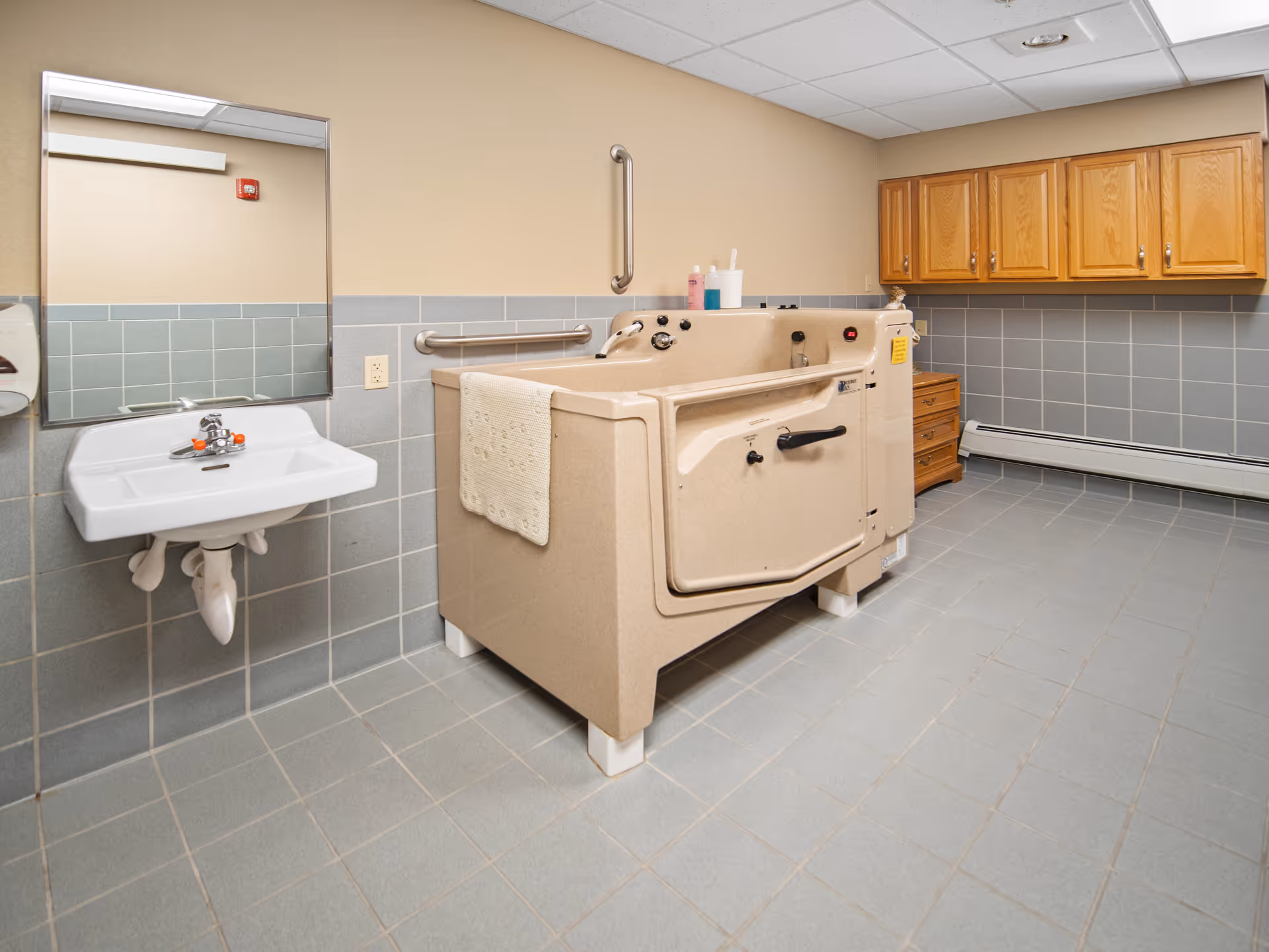 A bathroom with a beige assisted bathing tub, a white wall-mounted sink with a mirror above it, gray tiled walls and floor, wooden cabinets on the right wall, and grab bars installed near the tub.