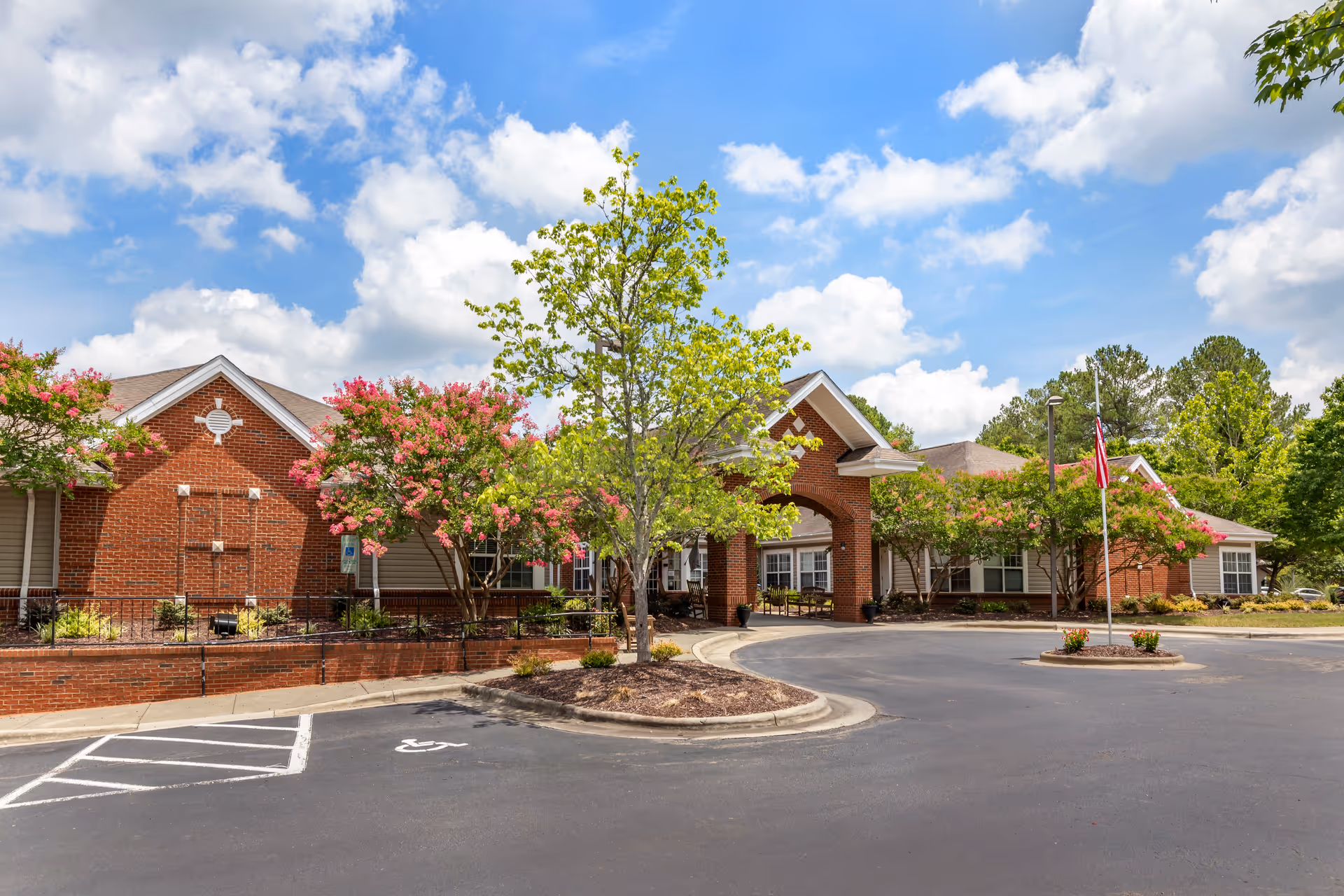 Exterior view of a brick building with a covered entrance, surrounded by trees with pink flowers and green foliage under a partly cloudy blue sky. There is a circular driveway with a flagpole displaying an American flag and a handicapped parking space visible in the foreground.
