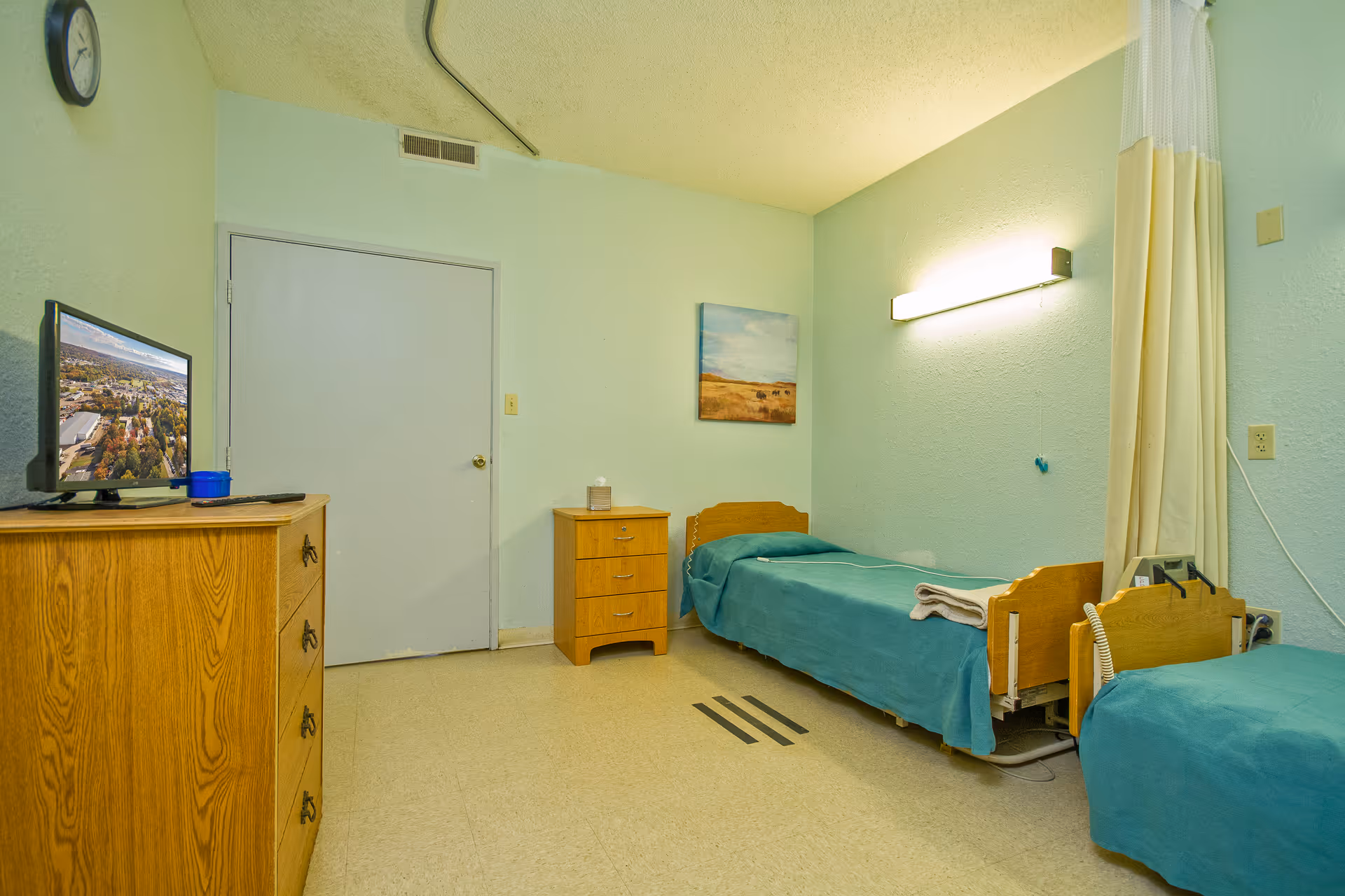 A simple room with two single beds covered with teal bedspreads, a wooden dresser with a TV on top, a small wooden nightstand with a tissue box, a wall-mounted light fixture, a clock, and a painting on the wall. The room has light green walls and a closed door.