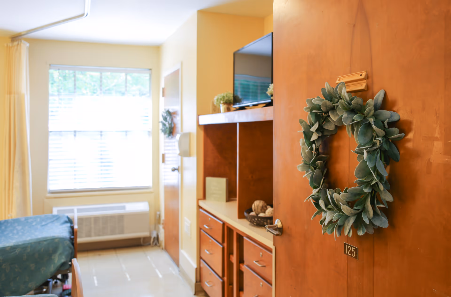 View into a senior living facility room with a wooden door decorated with a green leafy wreath and the number 125. Inside the room, there is a bed with a blue bedspread, a window with blinds, a heating unit below the window, and wooden cabinetry with a TV and decorative items on top.