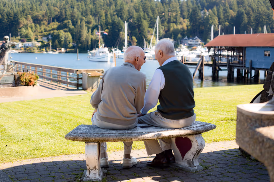 Two elderly men sitting on a stone bench with a heart design, facing a waterfront with boats docked and surrounded by trees, enjoying a sunny day outdoors.