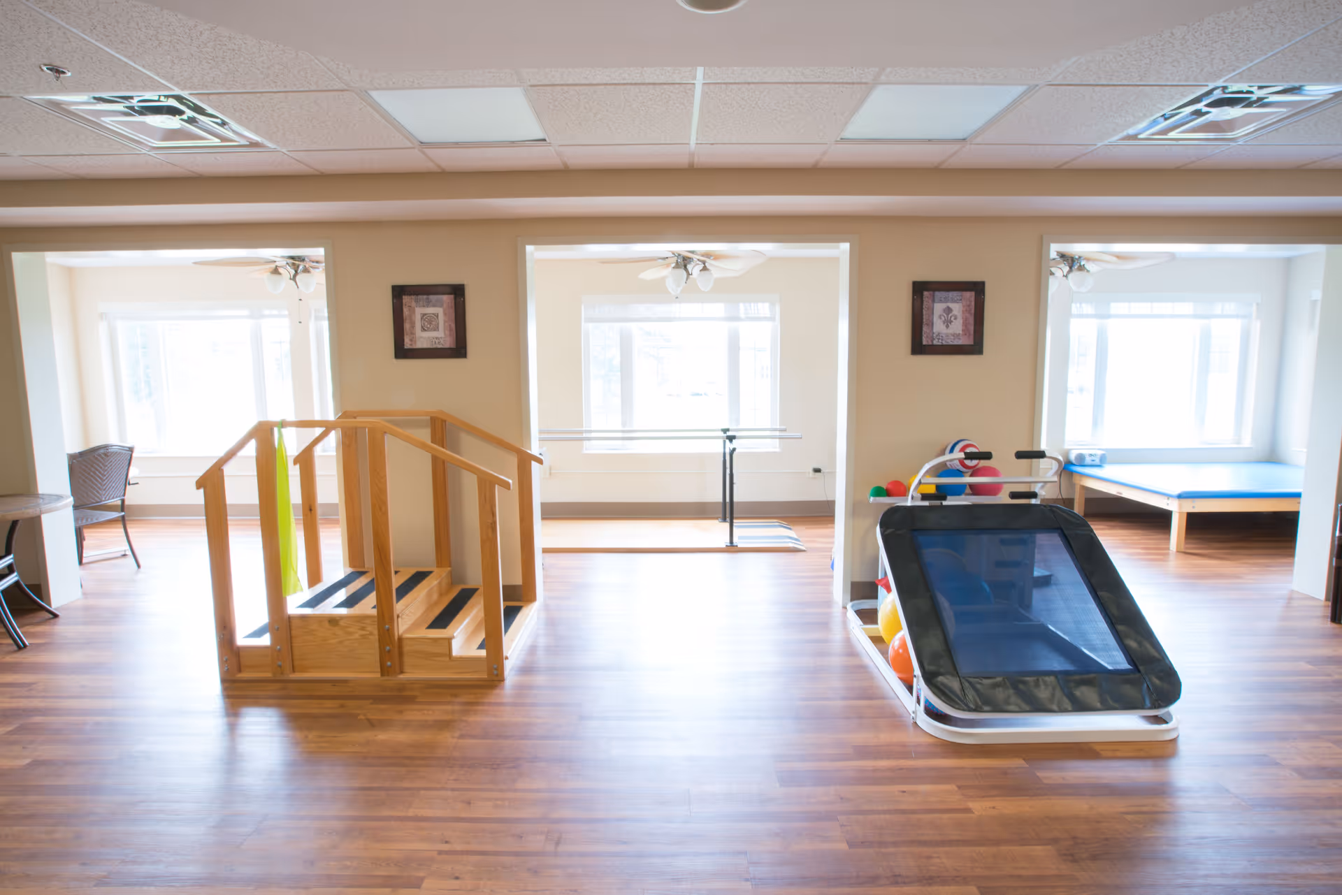 A bright room with wooden flooring featuring physical therapy equipment including a small wooden staircase with handrails, a rack holding colorful exercise balls, and a padded therapy table. The room has large windows allowing natural light and framed artwork on the walls.