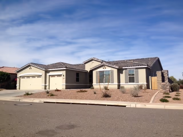 Single-story residential building with a beige exterior, tiled roof, and three garage doors. The front yard is landscaped with gravel and sparse desert plants. The sky is clear with some clouds.