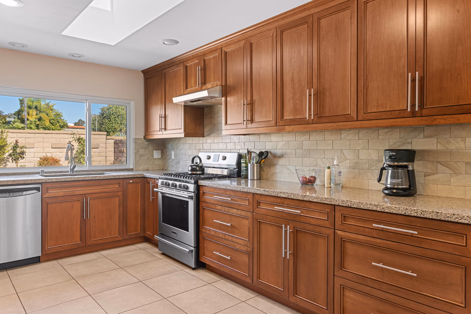 A modern kitchen with wooden cabinets, a stainless steel stove and dishwasher, a coffee maker on the counter, and a window showing an outdoor garden area with trees and a brick wall.