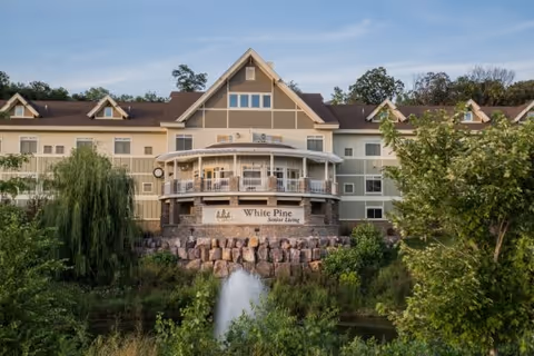 Exterior view of White Pine Senior Living building with a large porch, multiple windows, and a stone retaining wall. The foreground features greenery, trees, and a water fountain in a pond.