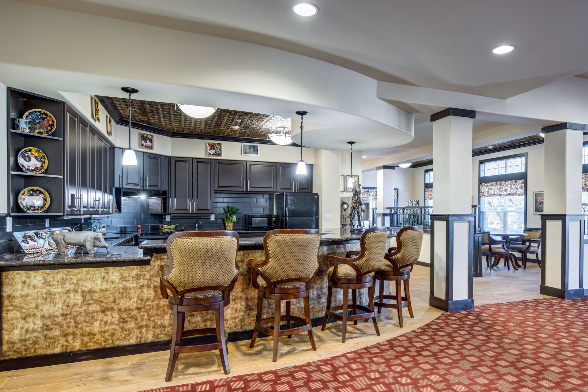 Interior view of a senior living facility kitchen and dining area. The kitchen features dark cabinetry, a black refrigerator, and a granite countertop with four upholstered bar stools. The space is well-lit with pendant lights and ceiling fixtures. Adjacent to the kitchen is a dining area with tables and chairs near large windows with patterned valances. The floor has a combination of wood and red patterned carpet, and the walls and columns are painted in neutral tones with dark trim.