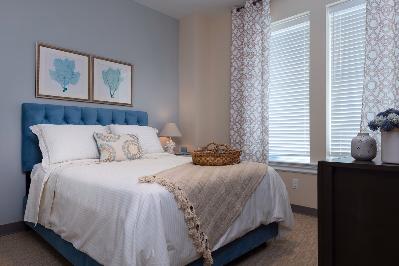 A neatly made bed with a blue upholstered headboard, white bedding, and decorative pillows. A beige throw blanket with tassels is draped across the foot of the bed with a woven basket on top. Two framed sea fan prints hang on the light gray wall above the bed. To the right, there are two windows with white blinds and patterned curtains. A dark wooden dresser with a vase of flowers and a decorative ceramic piece is visible in the foreground.