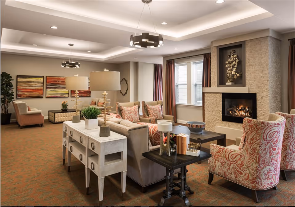 A cozy living room area in a senior living facility with a stone fireplace, beige and patterned armchairs, a beige sofa with patterned cushions, a white console table with two lamps and plants, a dark wooden coffee table with decorative items, and colorful abstract paintings on the wall. The room has recessed lighting and large windows with curtains.