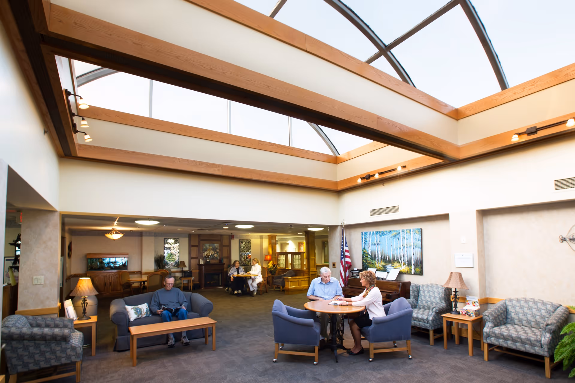 A bright and spacious common area in a nursing facility with a large skylight ceiling. Several elderly people are seated on sofas and chairs, some reading and others engaged in conversation. The room is furnished with comfortable seating, a coffee table, lamps, and a piano against the wall. There is an American flag and a large painting of a forest on the wall.