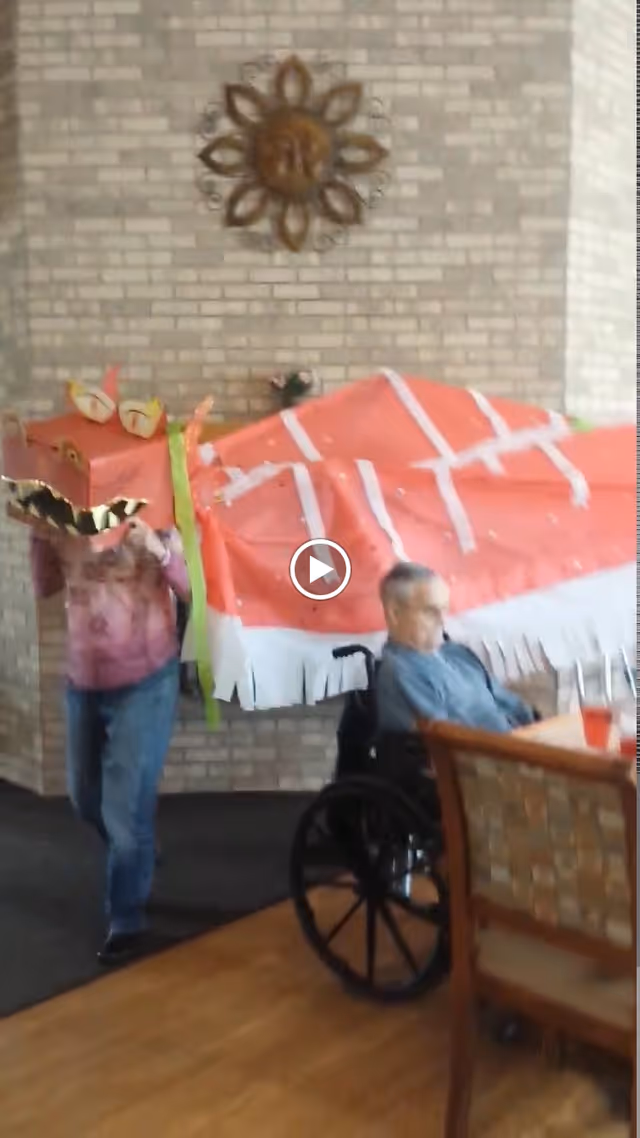 An elderly man in a wheelchair sitting at a table inside a room with brick walls. Another person is behind him, holding and wearing a colorful dragon costume, likely for a festive or cultural event. A decorative sun wall hanging is visible on the brick wall in the background.