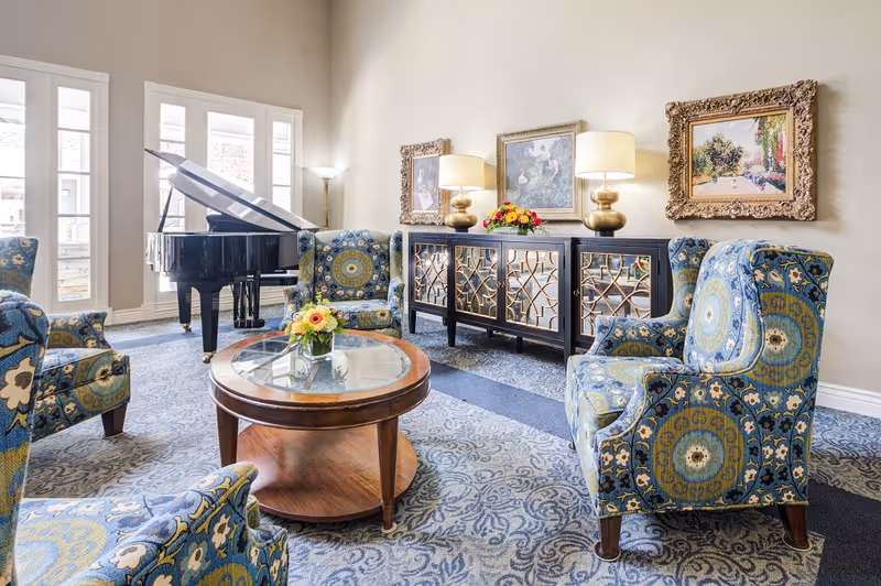 A bright and elegant living room area with patterned blue armchairs arranged around a round wooden coffee table with a glass top and a small flower arrangement. In the background, there is a black grand piano near tall windows with white shutters. Against the wall, a decorative black cabinet with gold geometric designs holds two gold lamps with white shades and a colorful flower arrangement. Three framed paintings hang on the beige wall above the cabinet.