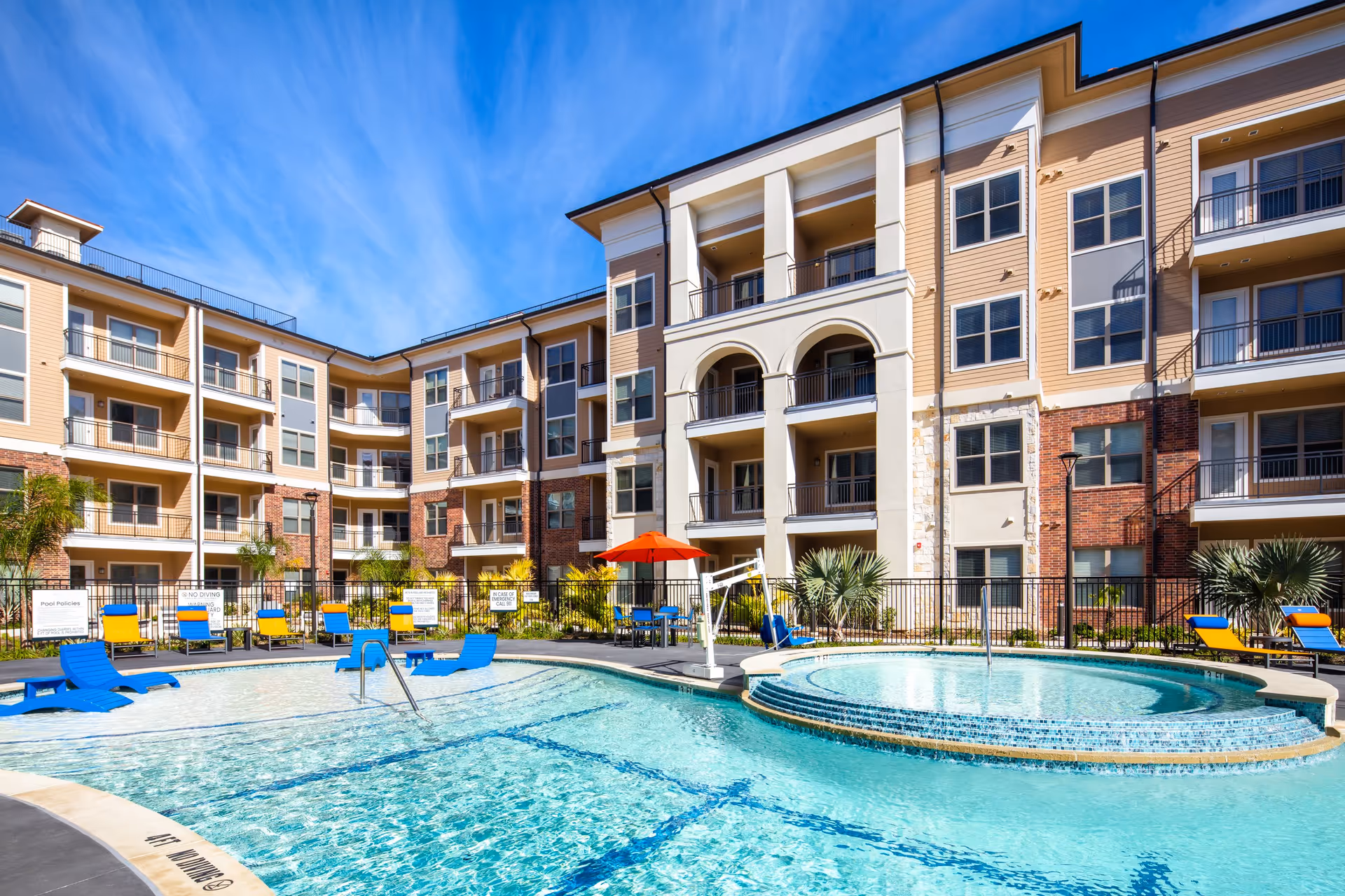 Outdoor swimming pool area at Arella on Jones Active Senior Community with blue water, poolside lounge chairs in blue and yellow, a round hot tub, and a multi-story residential building in the background under a clear blue sky.