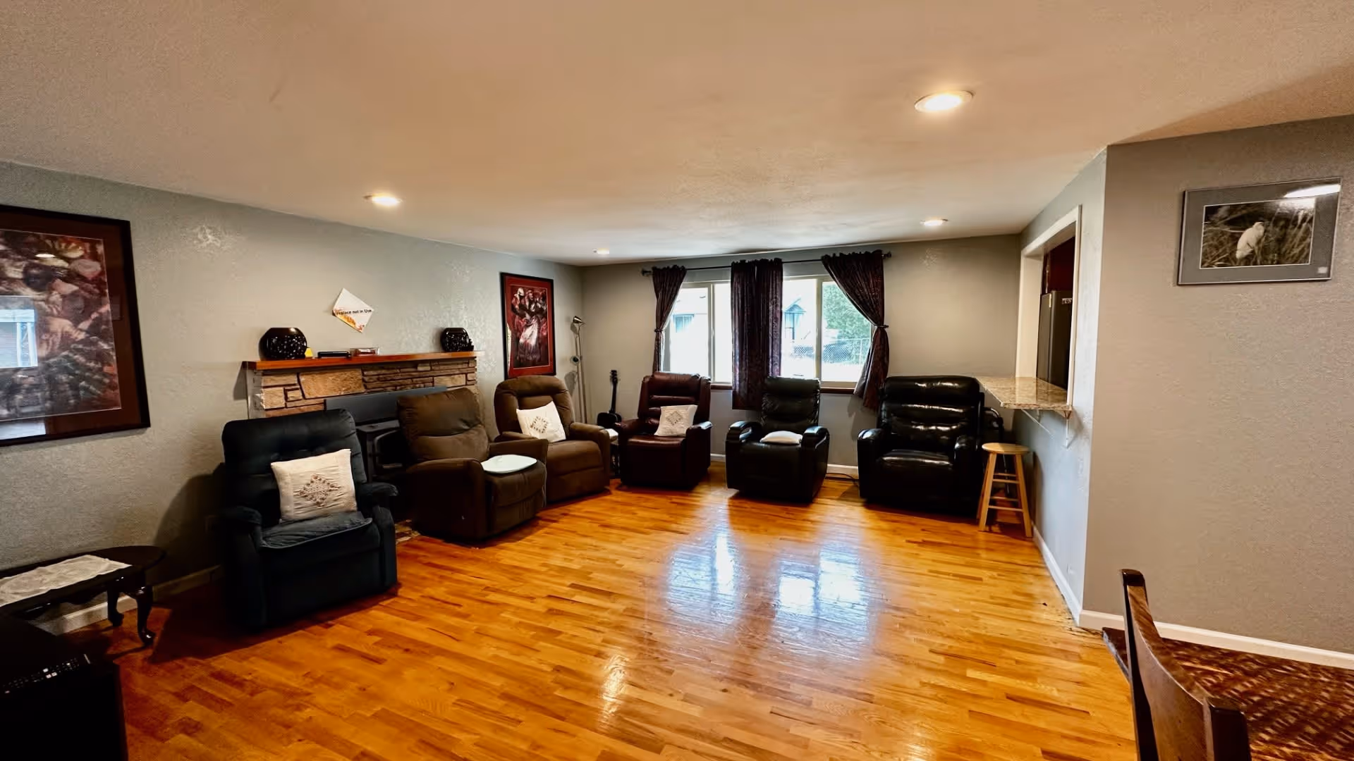 A spacious living room with five recliner chairs arranged in a semi-circle facing a stone fireplace. The room has hardwood floors, light gray walls, and recessed ceiling lights. Two windows with dark curtains allow natural light into the room. There are framed pictures on the walls and a small wooden stool near a pass-through counter to the kitchen.