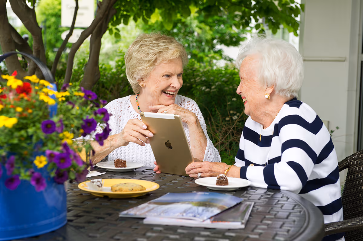 Two elderly women sitting at an outdoor table, smiling and looking at a tablet device together. There are plates with desserts and a cookie on the table, along with a blue pot of colorful flowers. The background shows green foliage and trees.