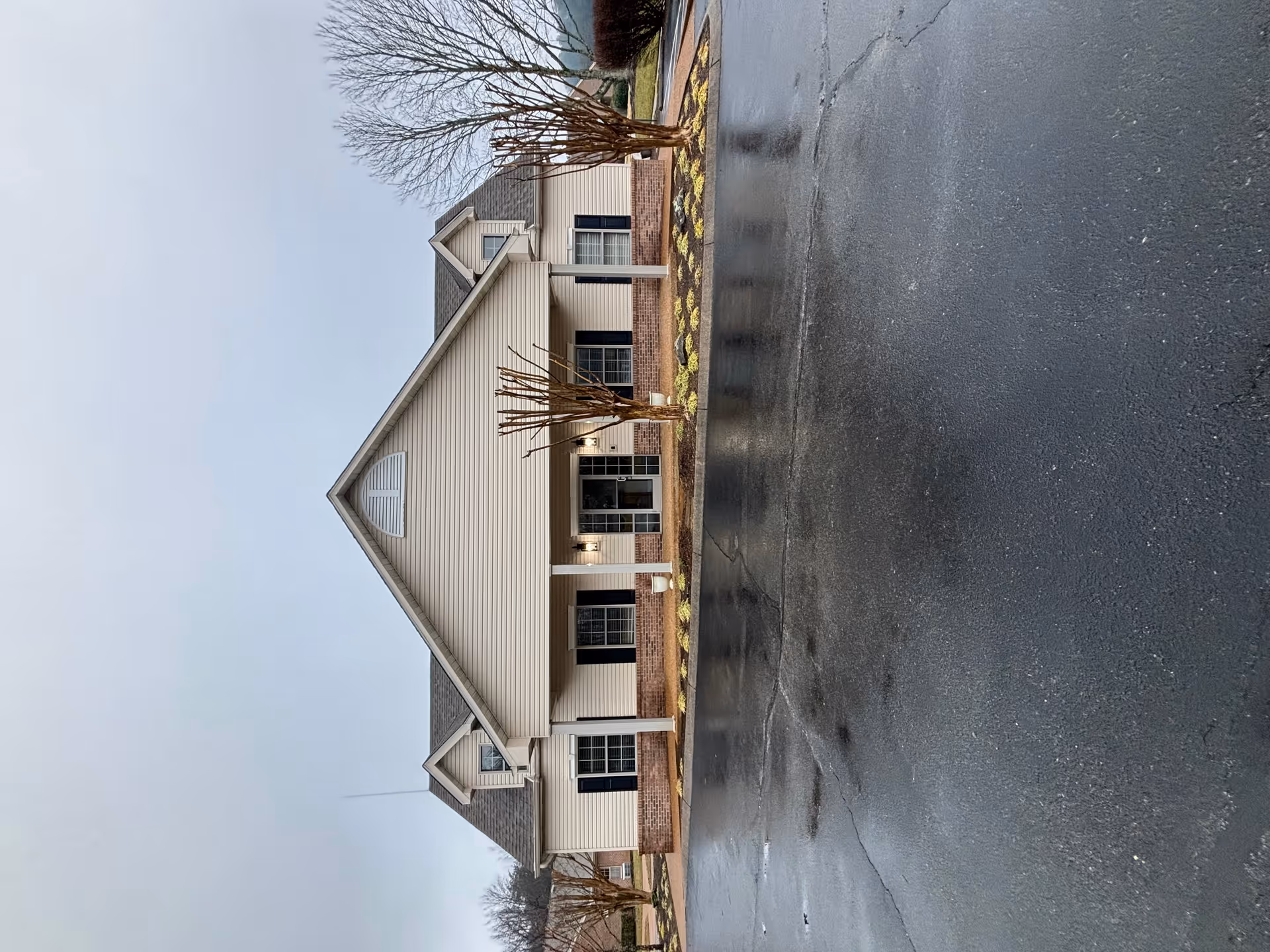 Front exterior view of a single-story building with beige siding and brick accents, featuring a covered entrance supported by white columns, two leafless trees in front, and a wet asphalt driveway under an overcast sky.