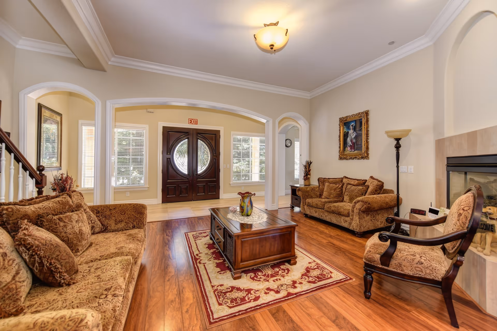 A spacious and well-lit living room with two patterned brown sofas, a matching armchair, a wooden coffee table on a decorative rug, a fireplace, and a painting on the wall. The room features wooden flooring, large windows with blinds, and a double-door entrance with an exit sign above it.