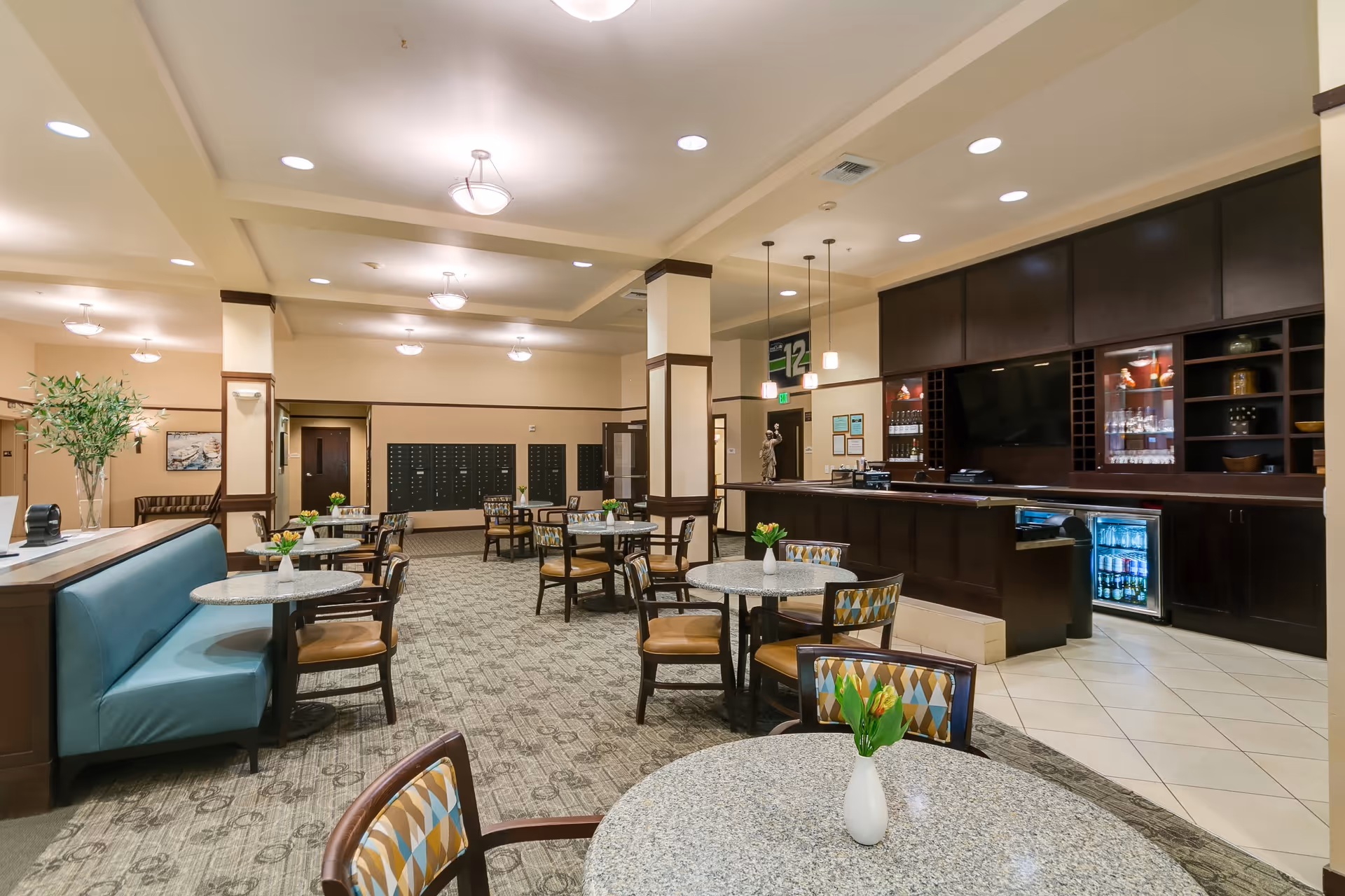 A spacious dining area in a senior living facility with round tables and chairs arranged neatly. Each table has a small white vase with yellow tulips. There is a blue cushioned bench along one side and a dark wood counter with cabinets and a small refrigerator in the background. The room is well-lit with ceiling lights and pendant lamps, and there are mailboxes along the far wall.