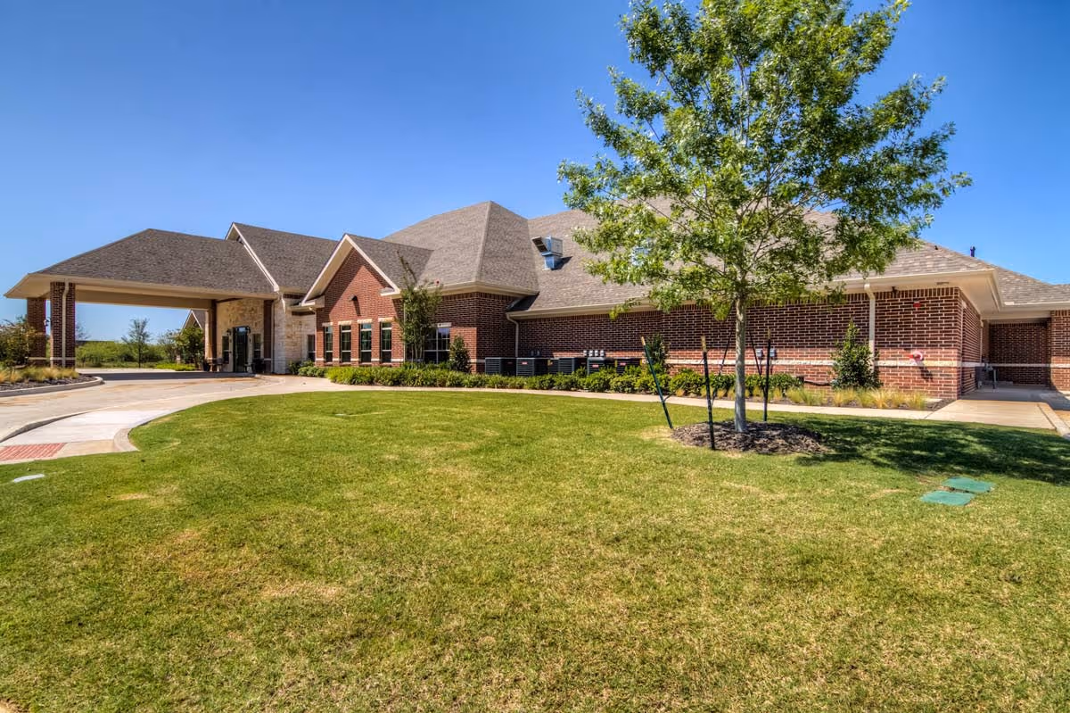 Exterior view of a single-story brick building with a covered entrance, surrounded by a well-maintained lawn and a tree under a clear blue sky.