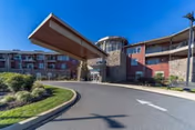 Exterior view of The Summit senior living facility showing a curved driveway leading to the main entrance with a large covered drop-off area. The building has multiple floors with balconies, stone and brick facade, and landscaped greenery under a clear blue sky.