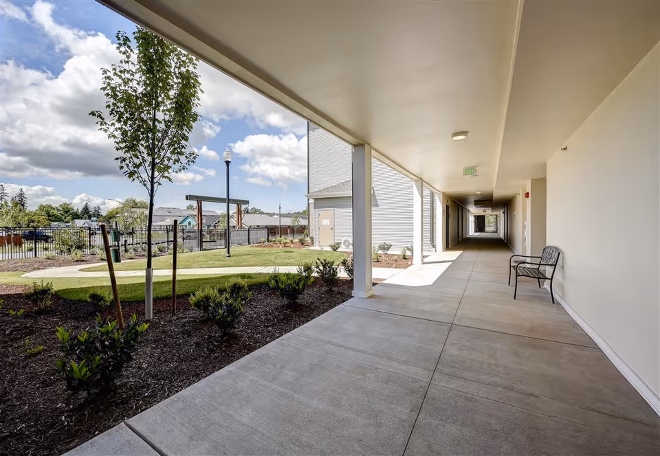 Covered outdoor walkway with a bench along the right side, leading to a garden area with small trees and shrubs. The sky is partly cloudy and the area is fenced with a view of nearby houses.