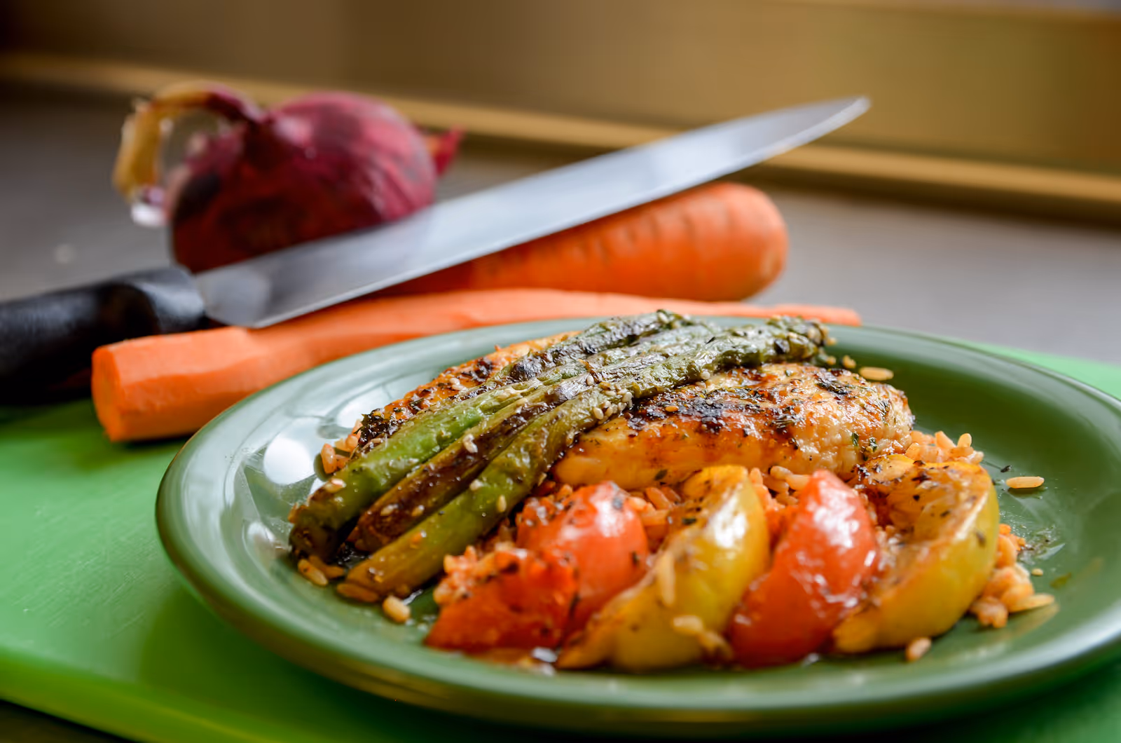 A green plate with grilled chicken breast topped with grilled asparagus, served on a bed of rice with roasted tomato and yellow bell pepper slices. In the background, there is a large kitchen knife resting on a carrot and a red onion on a green cutting board.