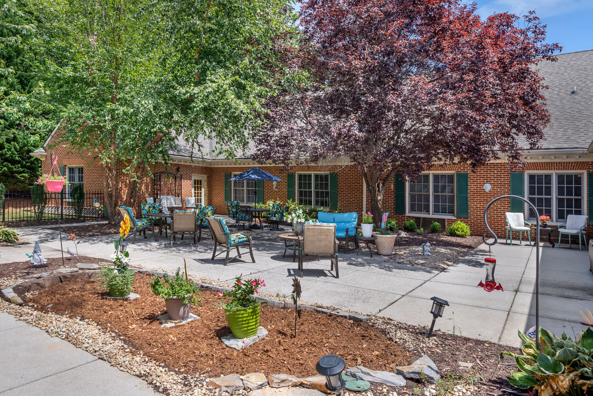 Outdoor patio area at Brookdale Midlothian with multiple seating arrangements including chairs and tables, some with cushions and umbrellas. The patio is surrounded by trees, potted plants, and garden beds with mulch and decorative stones. The building in the background is made of red brick with green shutters on the windows.