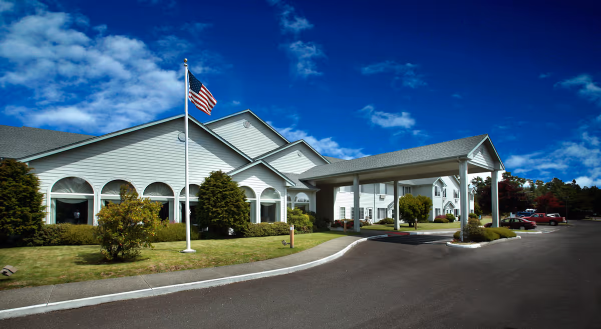 Exterior view of a single-story assisted living facility with white siding, a covered entrance, an American flag on a flagpole, and a parking area with cars under a partly cloudy blue sky.