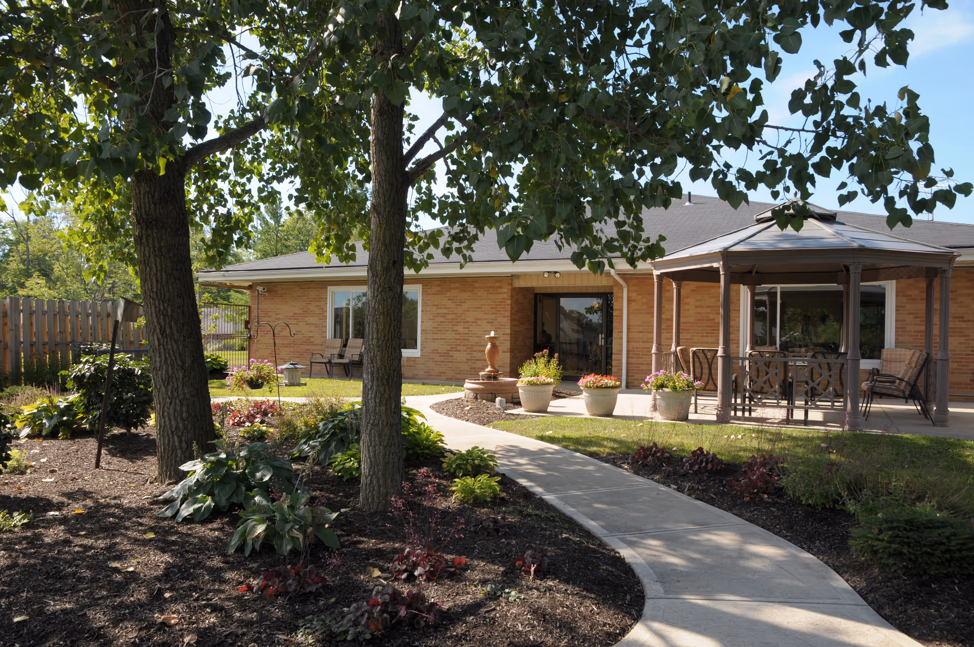 Landscaped courtyard with trees, flower beds, a curved walkway, a gazebo and the brick exterior of a senior living building.