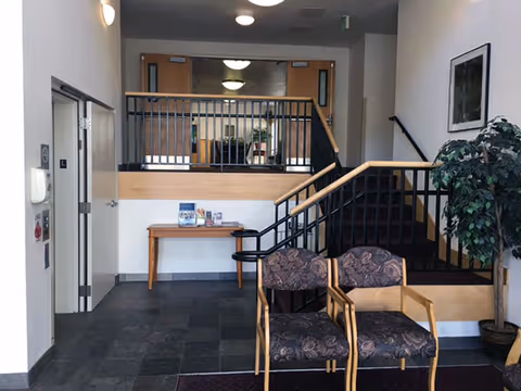Interior view of a senior living facility lobby area with two patterned armchairs in the foreground, a potted plant to the right, a wooden table with brochures against the back wall, and a staircase with black railings leading to an upper level.