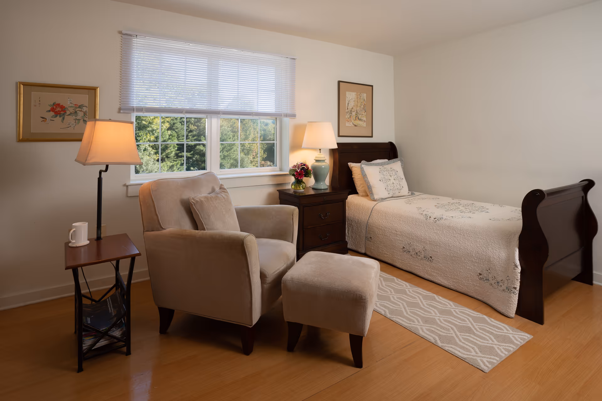 A cozy bedroom in a retirement community featuring a single dark wood bed with a white floral quilt and matching pillow. Next to the bed is a dark wood nightstand with a green lamp and a small flower arrangement. A beige armchair with a matching ottoman is positioned near a window with white blinds, accompanied by a small side table holding a white mug. The room has light-colored walls, wooden flooring, and framed artwork on the walls.