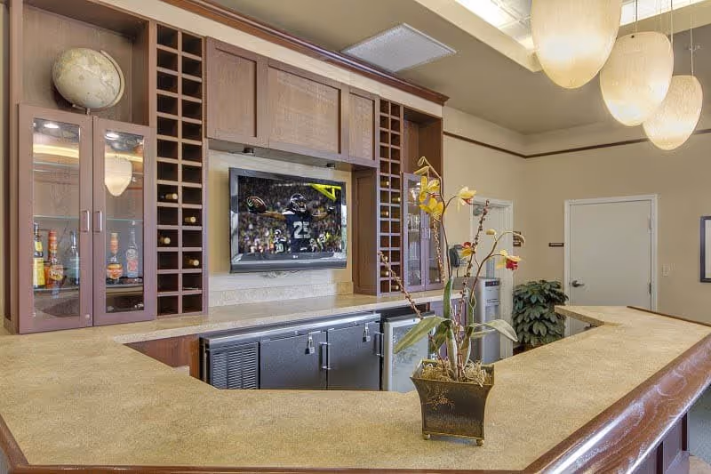 Interior view of a senior living facility's common area with a U-shaped counter bar. Behind the counter are wooden cabinets with glass doors displaying bottles, a mounted flat-screen TV showing a football game, and wine racks. A decorative plant is placed on the counter, and three large pendant lights hang from the ceiling. There is a door and a plant in the background.