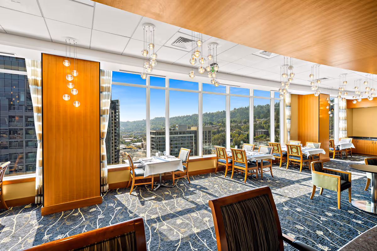 Sunlit dining room with tables and chairs beside large floor-to-ceiling windows overlooking the city and tree-covered hills.
