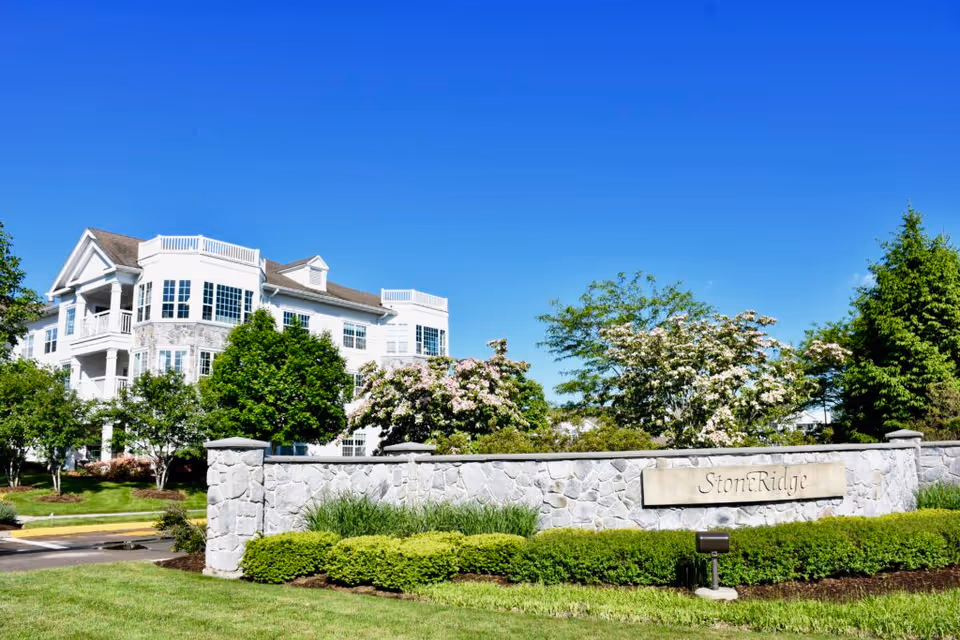 Exterior view of StoneRidge Senior Living facility with a large white multi-story building surrounded by green trees and bushes under a clear blue sky. A stone wall with a sign reading 'StoneRidge' is in the foreground.