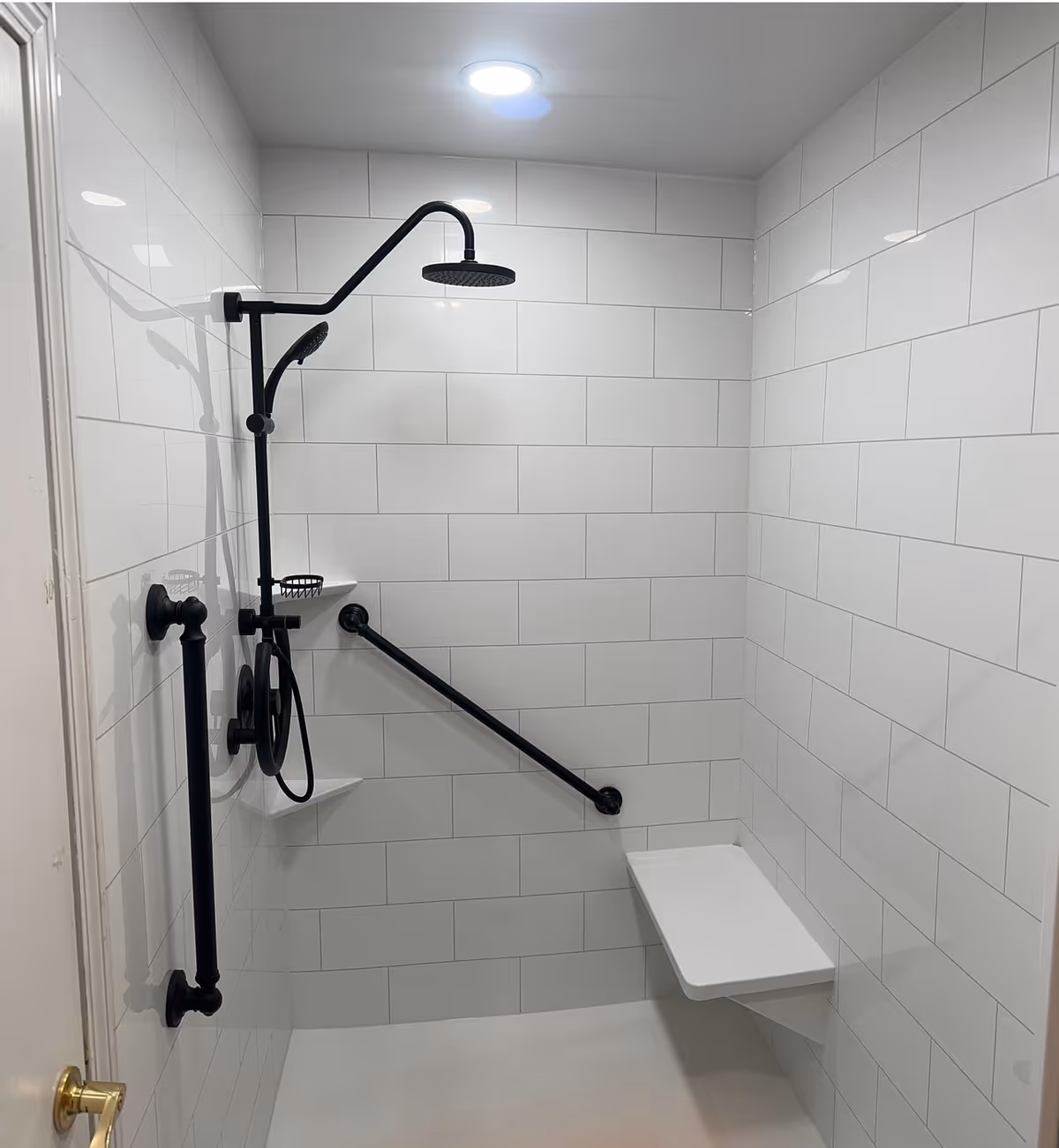 A modern shower area with white tiled walls, featuring a black rain showerhead, a handheld shower, two black grab bars, a small corner shelf, and a white foldable shower seat.